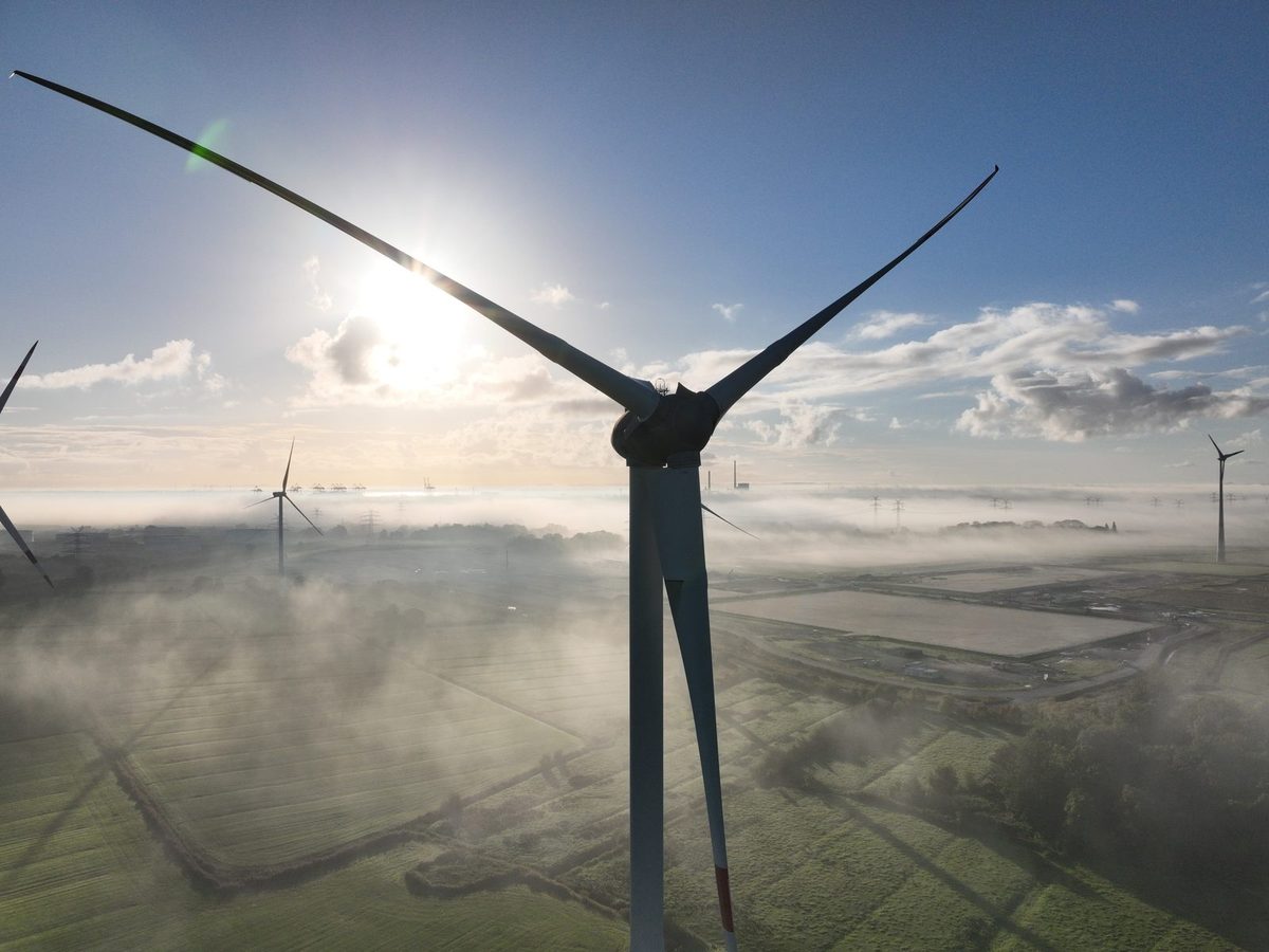 Windenergieanlage aus der Luft auf Nabenhöhe, Rotor im Stillstand in Y-Stellung mit einem Blatt nach unten und roter Blattspitzenmarkierung, Sonne hinter der Anlage, Bodennebel über den Feldern, weitere Anlagen im Hintergrund