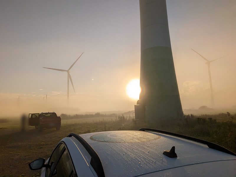 Sonnenaufgang im Nebel zwischen Windenergieanlagen, im Vordergrund das Dach eines Einsatzfahrzeugs